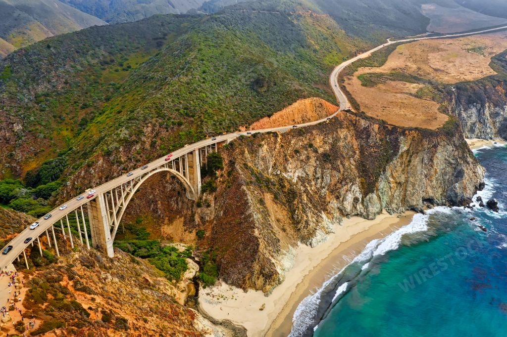 A beautiful aerial shot of green hills and a curvy narrow bridge going along the cliffs with the sea on the side