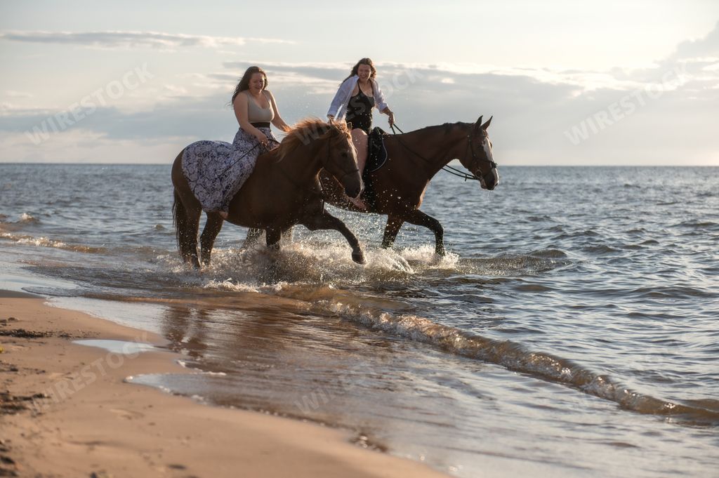 Two women on horseback riding in the ocean at sunset