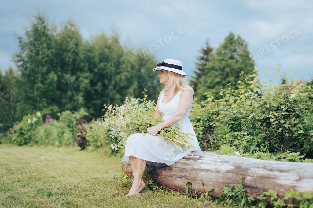 A beautiful blonde woman in a white dress and a hat holding flowers and sitting on a tree trunk