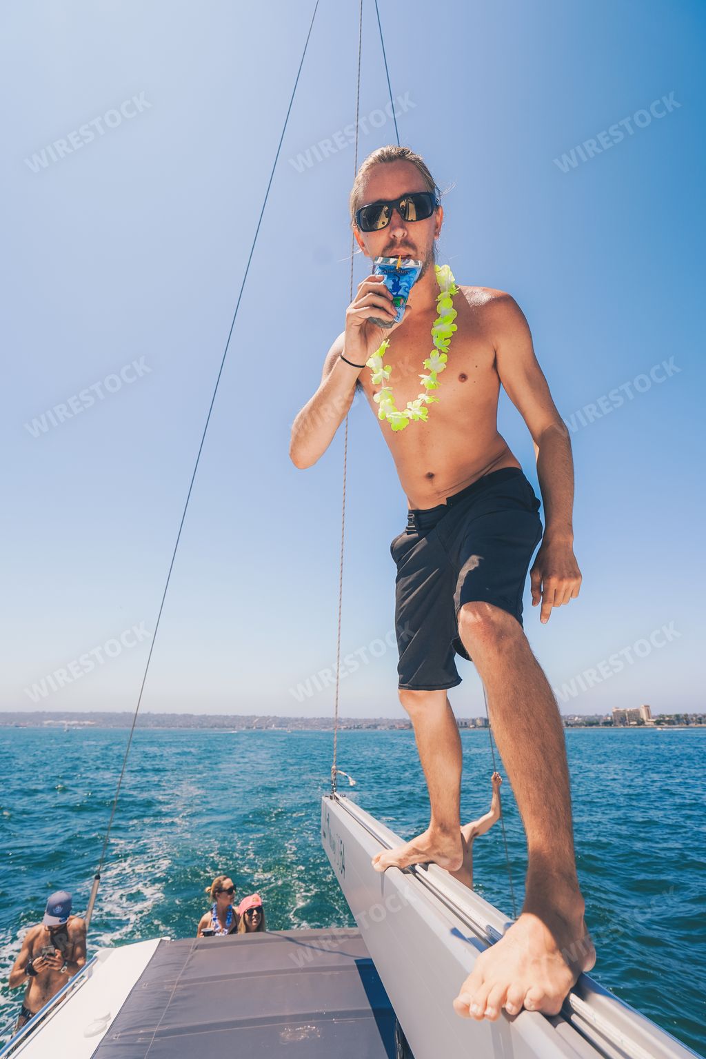 A vertical shot of a male with sunglasses standing on the edge of a white ship and drinking juice on a sunny day