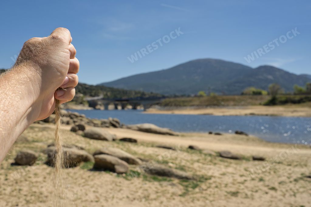 A man's hand pouring sand onto a shoreline in front of a tranquil lake, the drying out reservoir