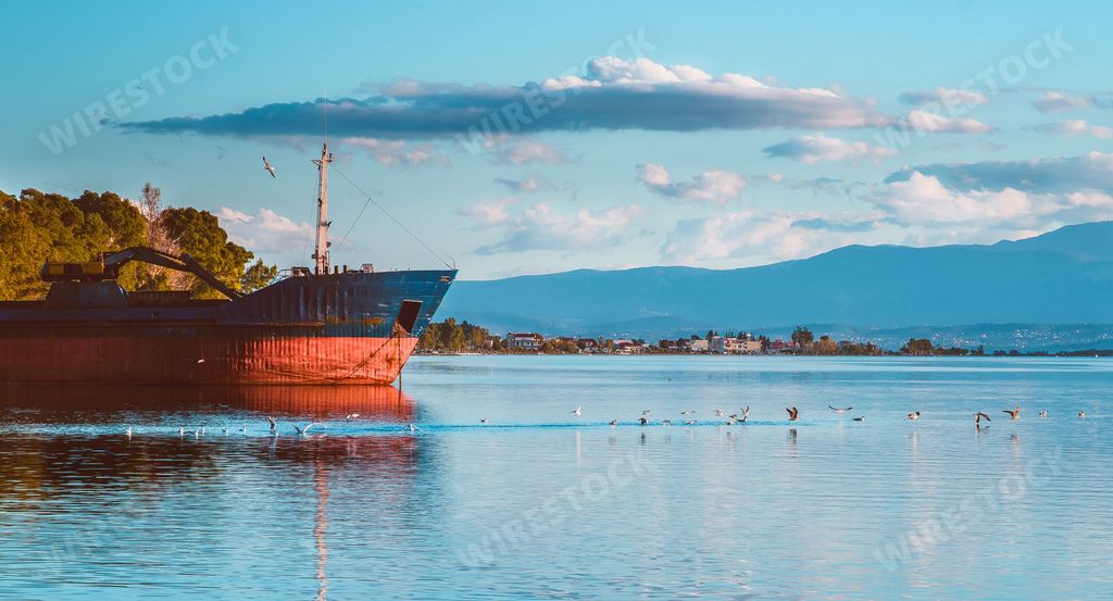 A scenic shot of a lake with an abandoned ship on it, while birds fly over the lake water on a sunny day