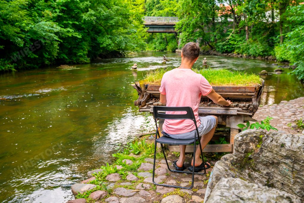 A young male playing an abandoned grand piano covered in the grass near the Vilnia River, Vilnius, Lithuania