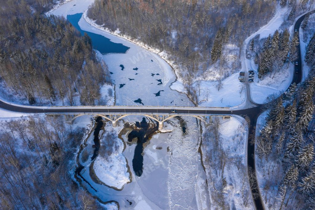 An aerial shot of a curvy road over a flowing river through the snow-covered forest