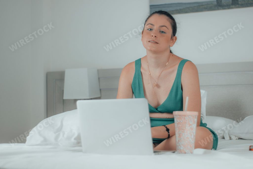 A closeup of a female sitting on the bed with crossed legs,a computer in front of, bottle cup next to it, looking at the camera