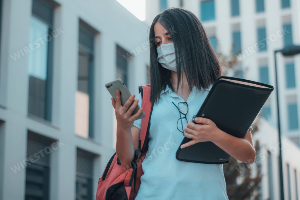 A low angle shot of a white Caucasian woman looking on her phone with a backpack and drawing kit wearing a face mask.