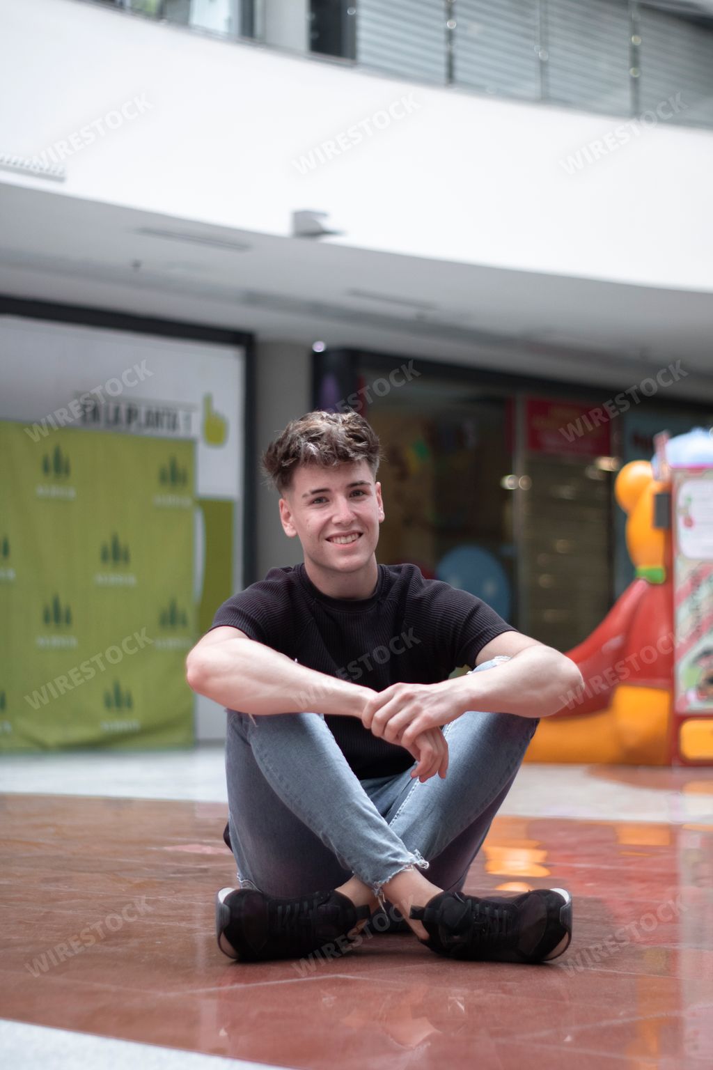 A vertical shot of an attractive Caucasian man sitting on the floor of a mall, smiling
