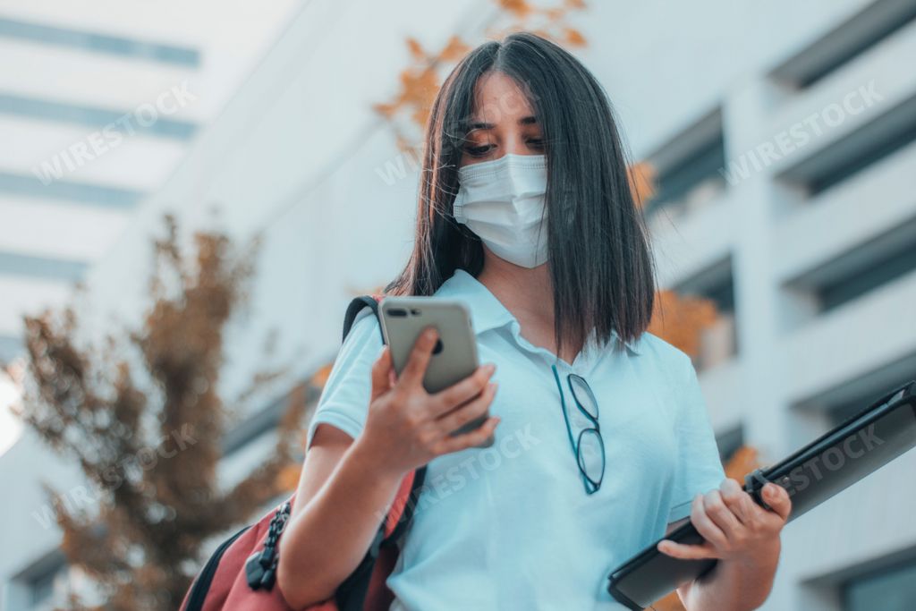 Low angle shot of a woman looking on her phone with a backpack and drawing kit wearing a face mask.