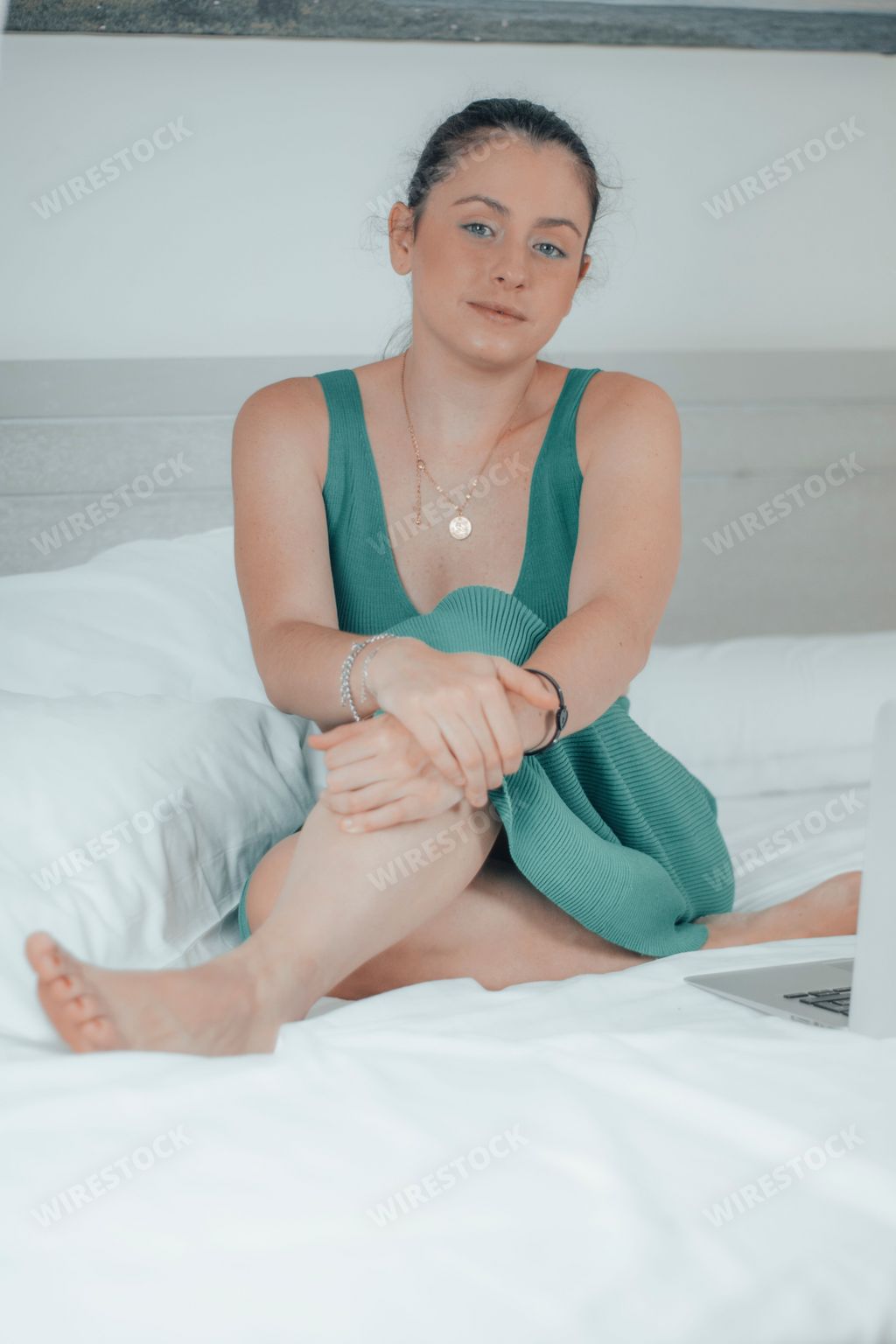 A vertical closeup of a female sitting on the bed with crossed legs, looking at the camera, white bedding blurred background