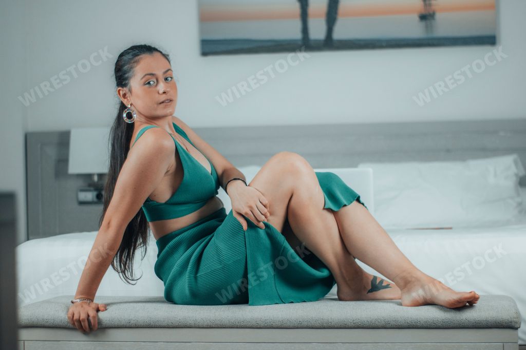 A side closeup of a female sitting on the bed with crossed legs, white bedding blurred background