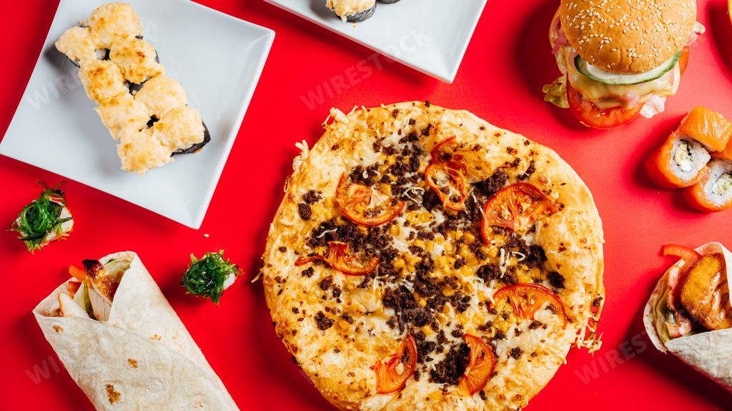 A top view of fast food delicacies including pizza, burger, wraps and sushi rolls on a red table