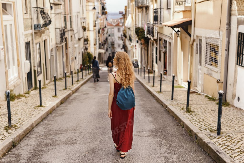 A young female tourist in a red dress walking through a road surrounded by buildings under the sunlight