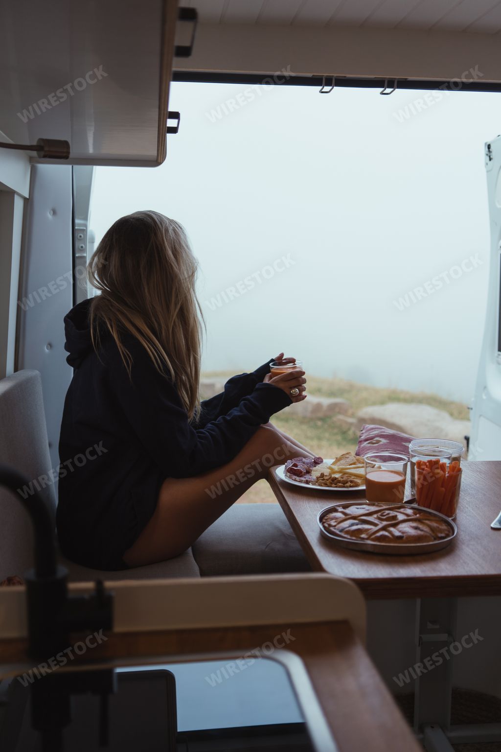 A vertical photo of a woman sitting by a window, watching nature and eating lunch