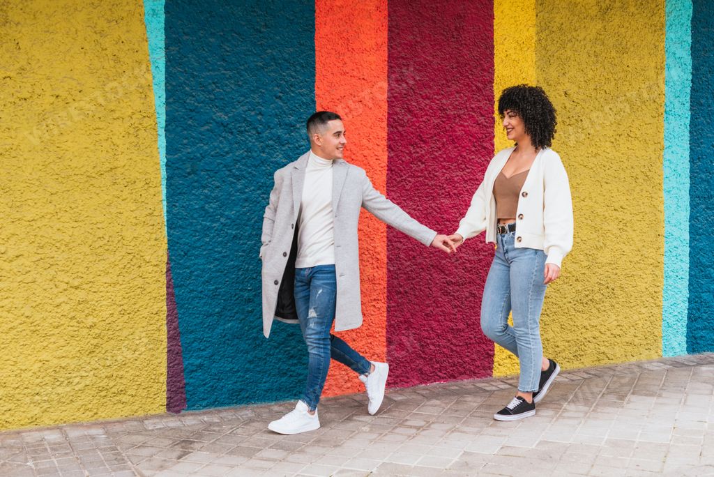 A young happy couple holding hands and walking through a pavement against a colorful wall