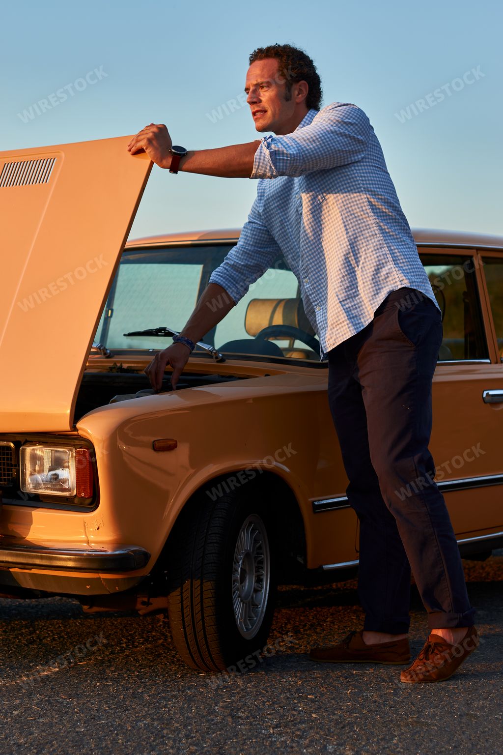 A vertical shot of a male model standing by his old vintage car with front side hood open