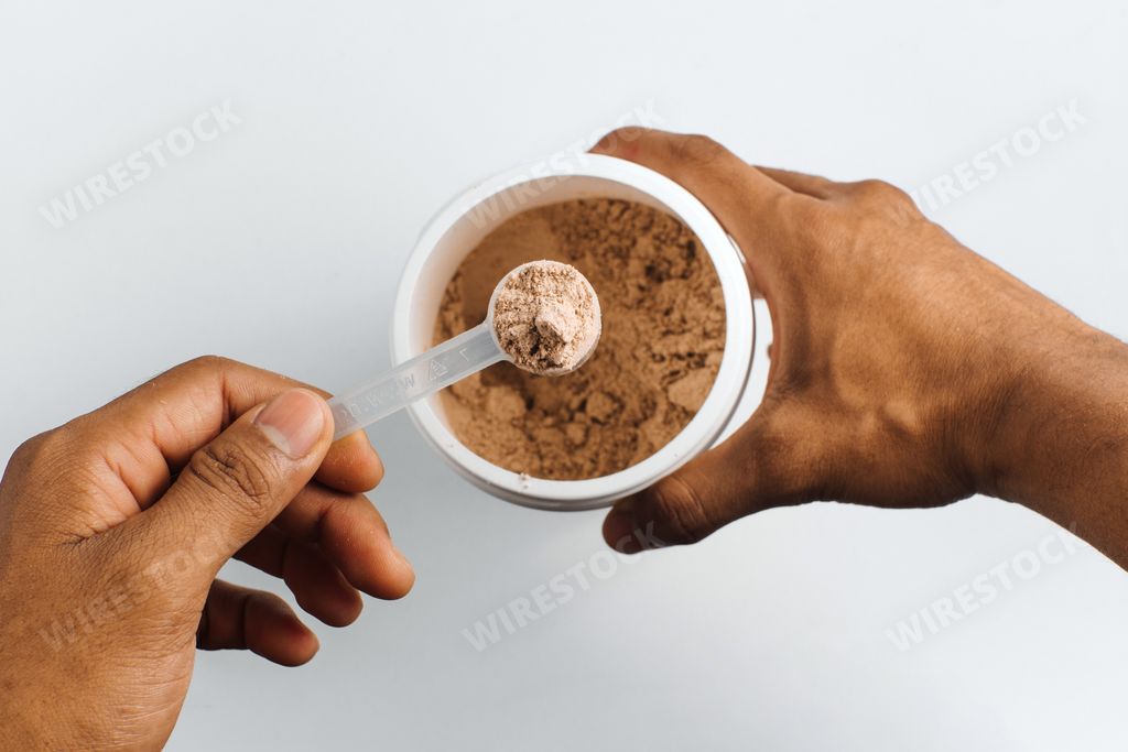 A person's hand holding a measuring spoon with chocolate protein powder over white background