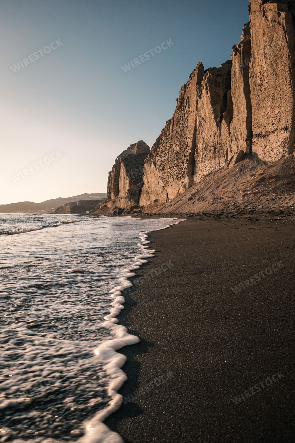 A gorgeous vertical shot of the Eros Beach in Santorini, Greece with a low frothy tide at the shore overlooked by proud cliffs