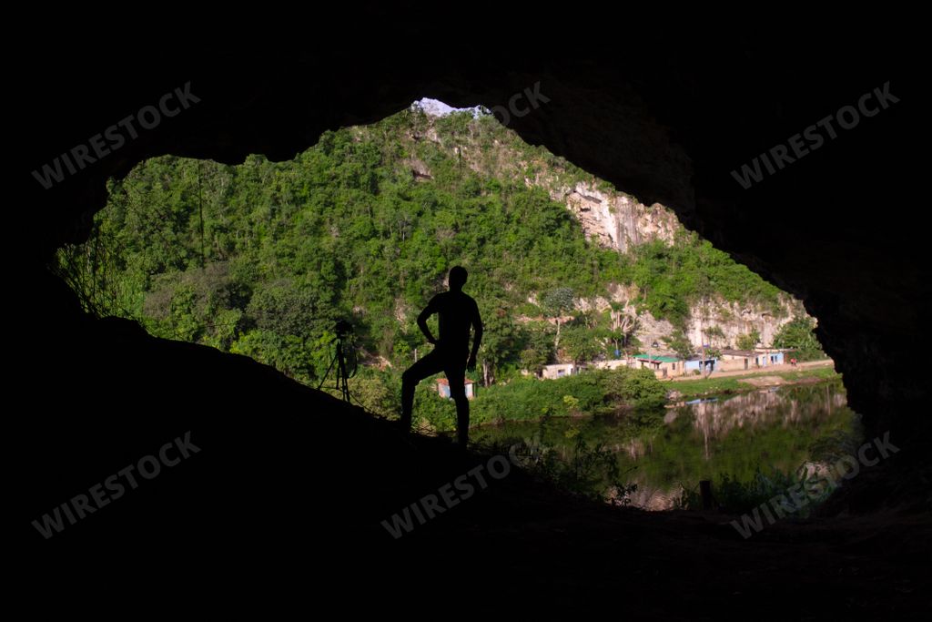 A Silhouette view of A Hispanic man standing inside a dark cave looking at green hills