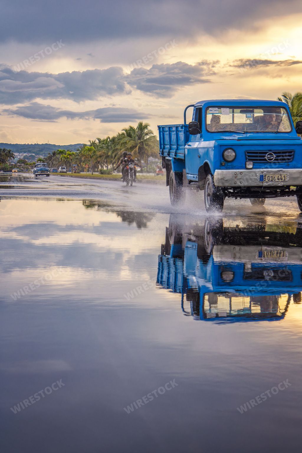 Two male adults driving a classic blue pickup truck through a body of water, creating a large splash with the wheels
