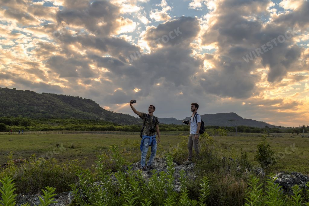 A beautiful scene of two Hispanic males at large grassland taking a selfie with mountains and a sunset sky