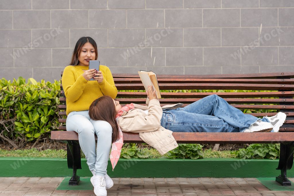 Two latina women sitting and lying on a bench while reading a book and using the mobile