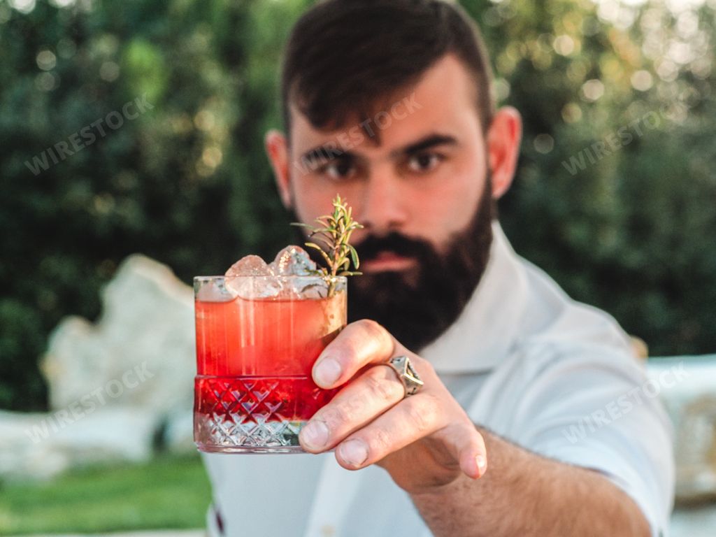 A young male with a beard showing a glass of Conte speziato cocktail standing outside