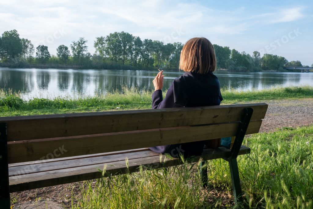 adult mature Woman smoking with her legs crossed sitting on a park bench near a lake in springtime