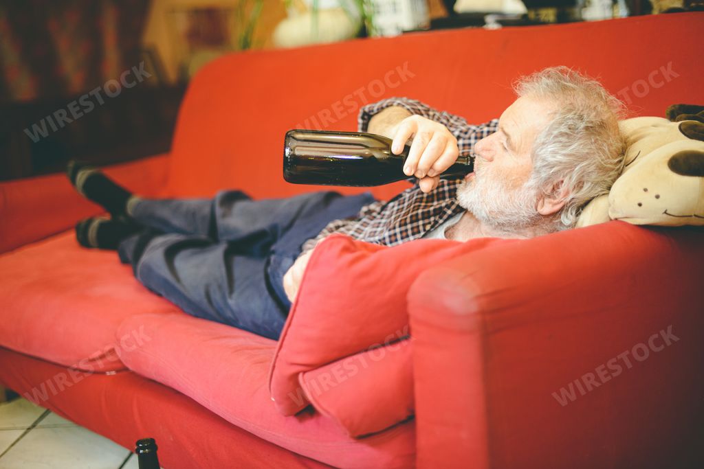 A shot of a Caucasian adult man laying on a couch and drinking some wine from a bottle.