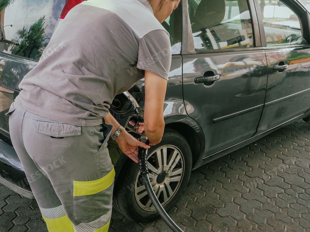 A man is seen sitting in the driver's seat of a car at a gas station, likely filling up the tank