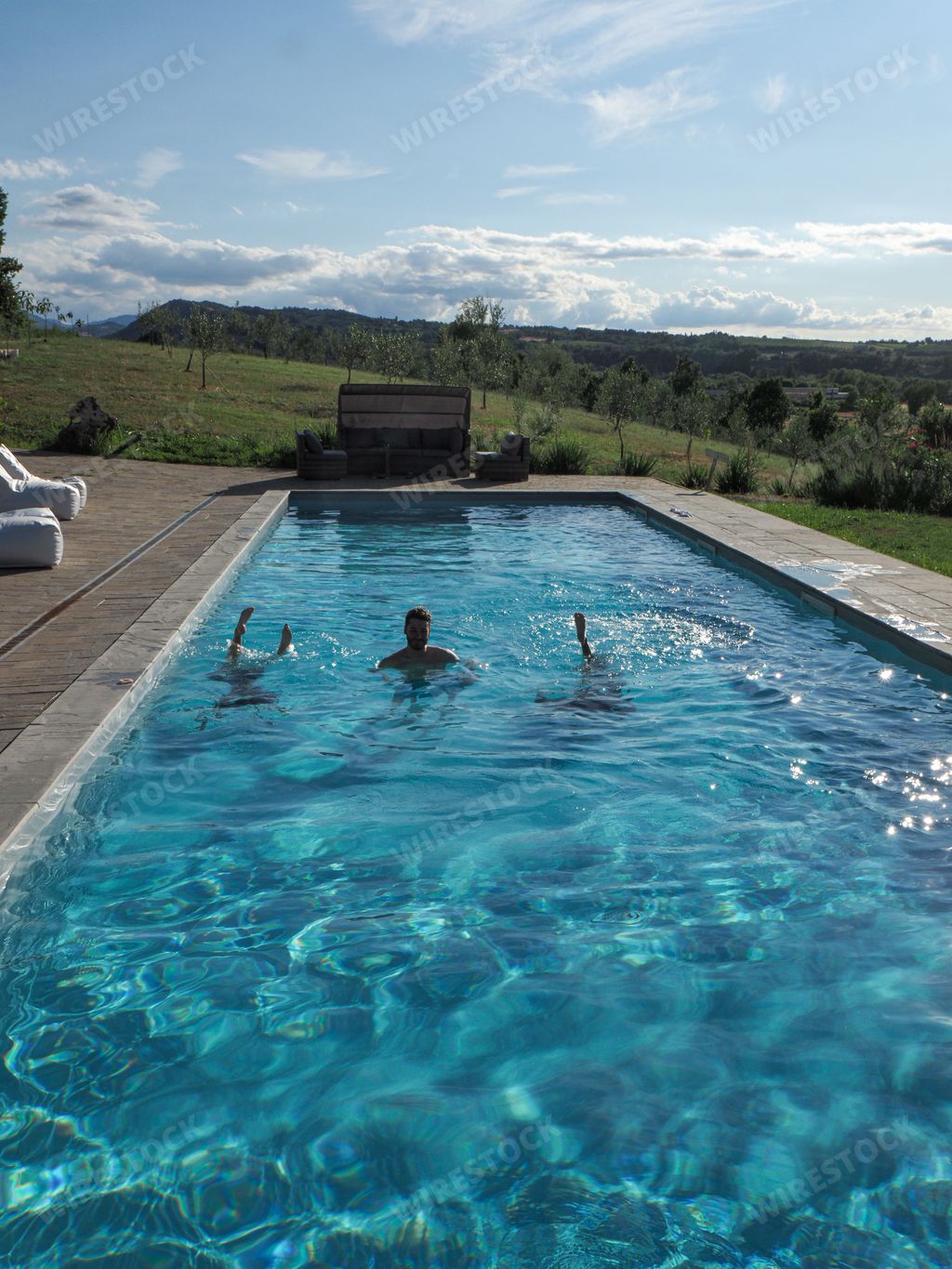 A vertical shot of a Caucasian man in the pool with two upside-down women, on a sunny day