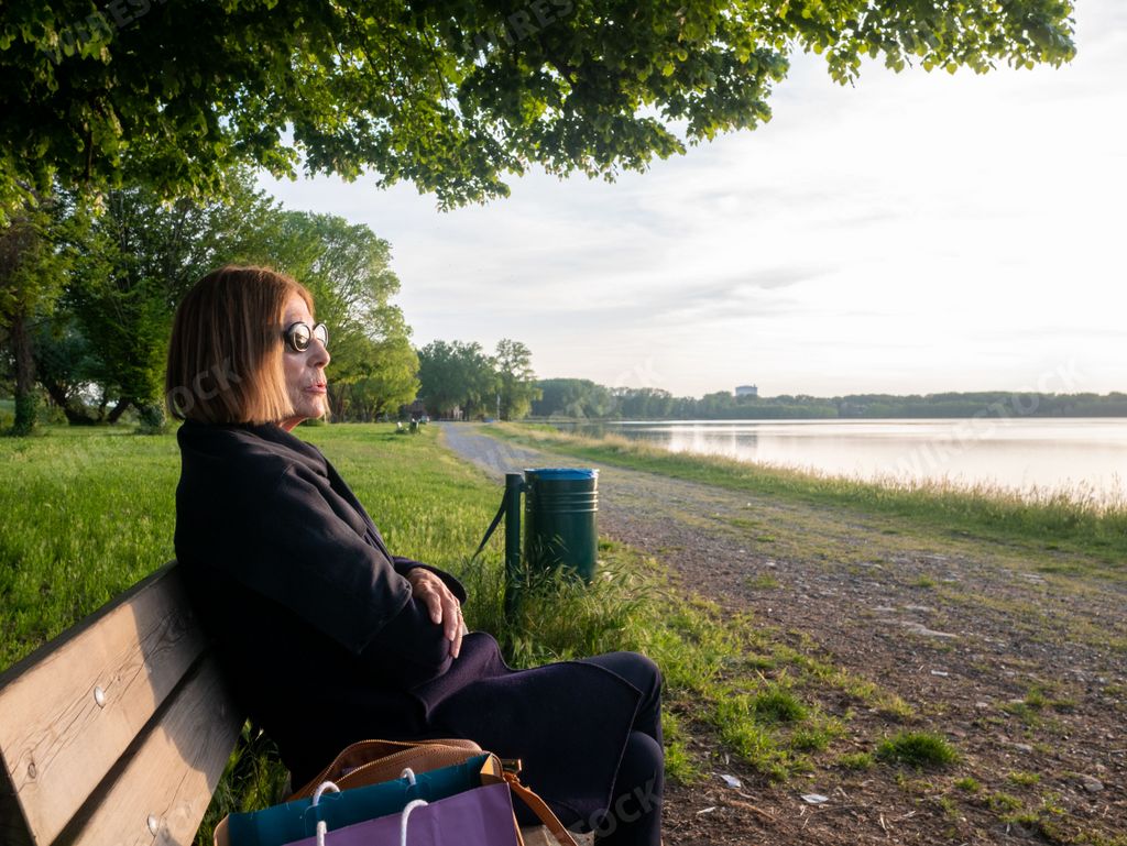 adult mature Woman relaxing with her legs crossed sitting on a park bench near a lake in springtime