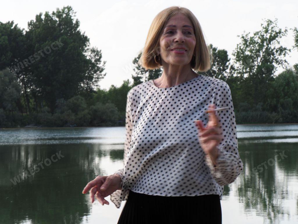 A joyful senior Caucasian woman smoking a cigarette in the park near the lake