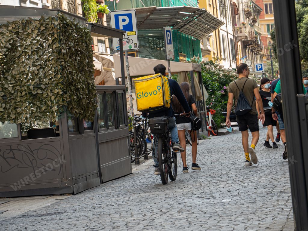 A "Glovo" food delivery man riding a bicycle on busy street in Milan, Italy