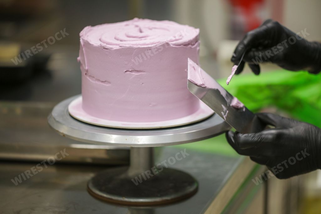A person dressed in black gloves meticulously slices into a creamy pink-frosted cake, making precise cuts