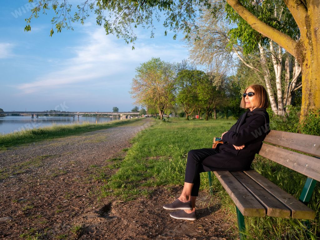 adult mature Woman relaxing with her legs crossed sitting on a park bench near a lake in springtime