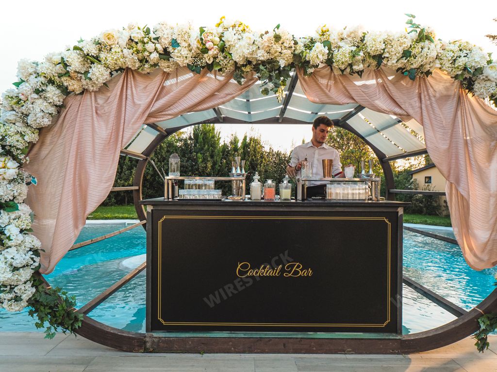 A Caucasian male bartender preparing a mai tai cocktail beahing the bar table outdoors near a pool