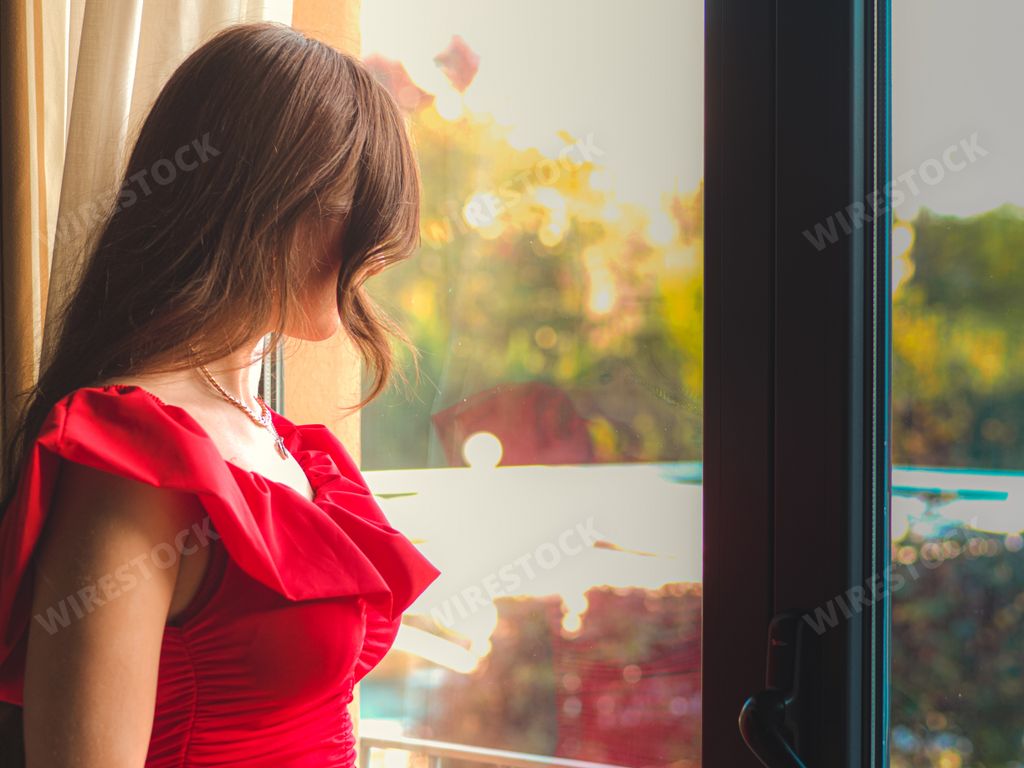 A Caucasian brunette girl with a red dress on valentine day  standing next to a glass balcony at home
