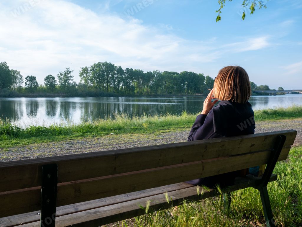 adult mature Woman smoking with her legs crossed sitting on a park bench near a lake in springtime