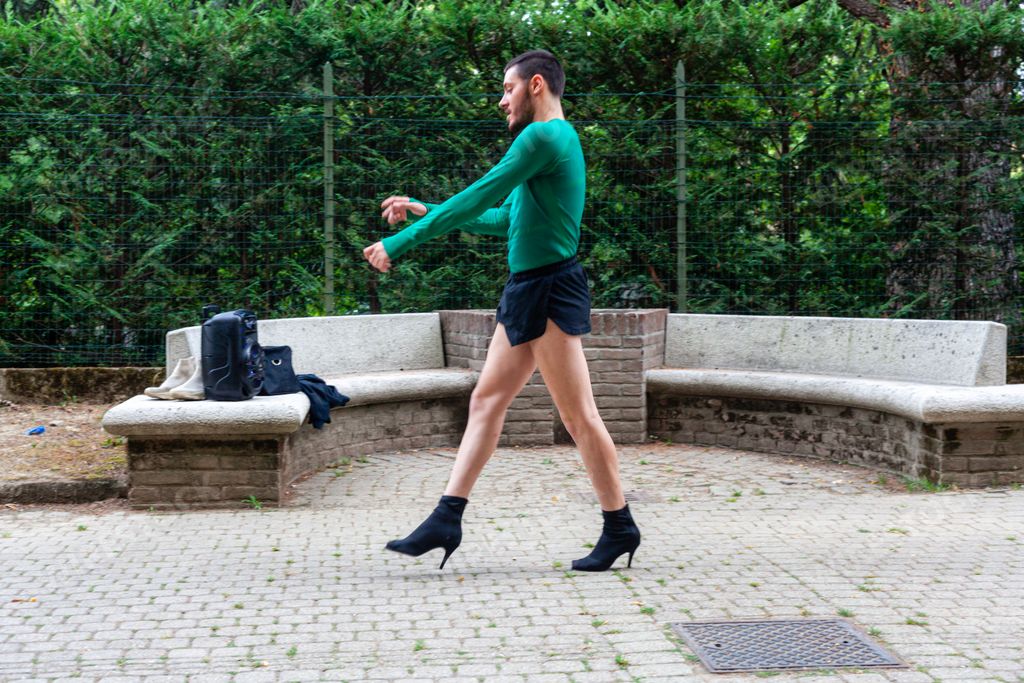 A young Caucasian man wearing high heels, shorts and a green shirt while posing in a park