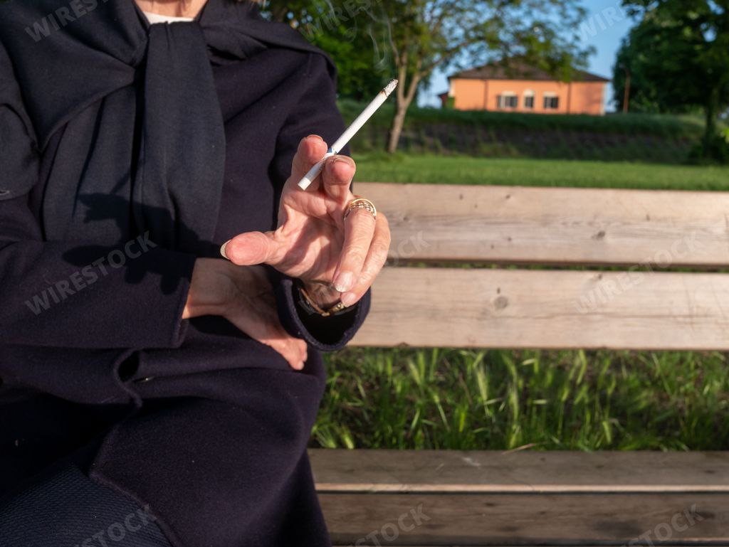 adult mature Woman smoking with her legs crossed sitting on a park bench near a lake in springtime