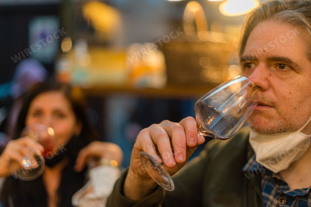 A middle-aged male with a face mask pulled down to his chin is drinking wine in the pub with friends