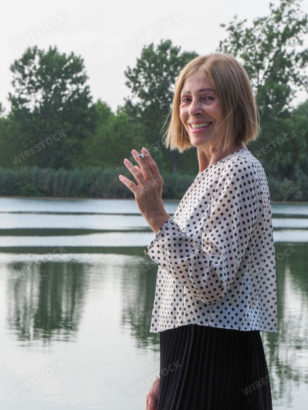 A joyful elderly woman in a white blouse with dots smoking a cigarette in a park near the water, aging free and wellness concept