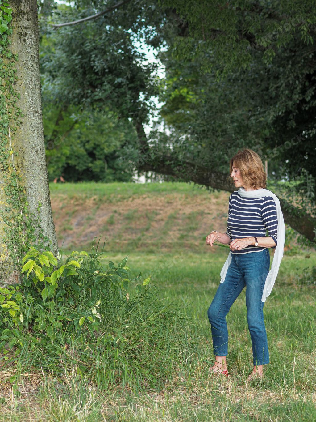 A Caucasian senior woman wearing a striped shirt and denim pants in a park