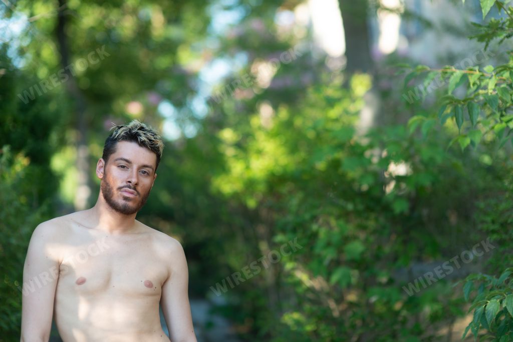 A young half-naked man with blonde dyed hair looking straight to the camera in the forest.