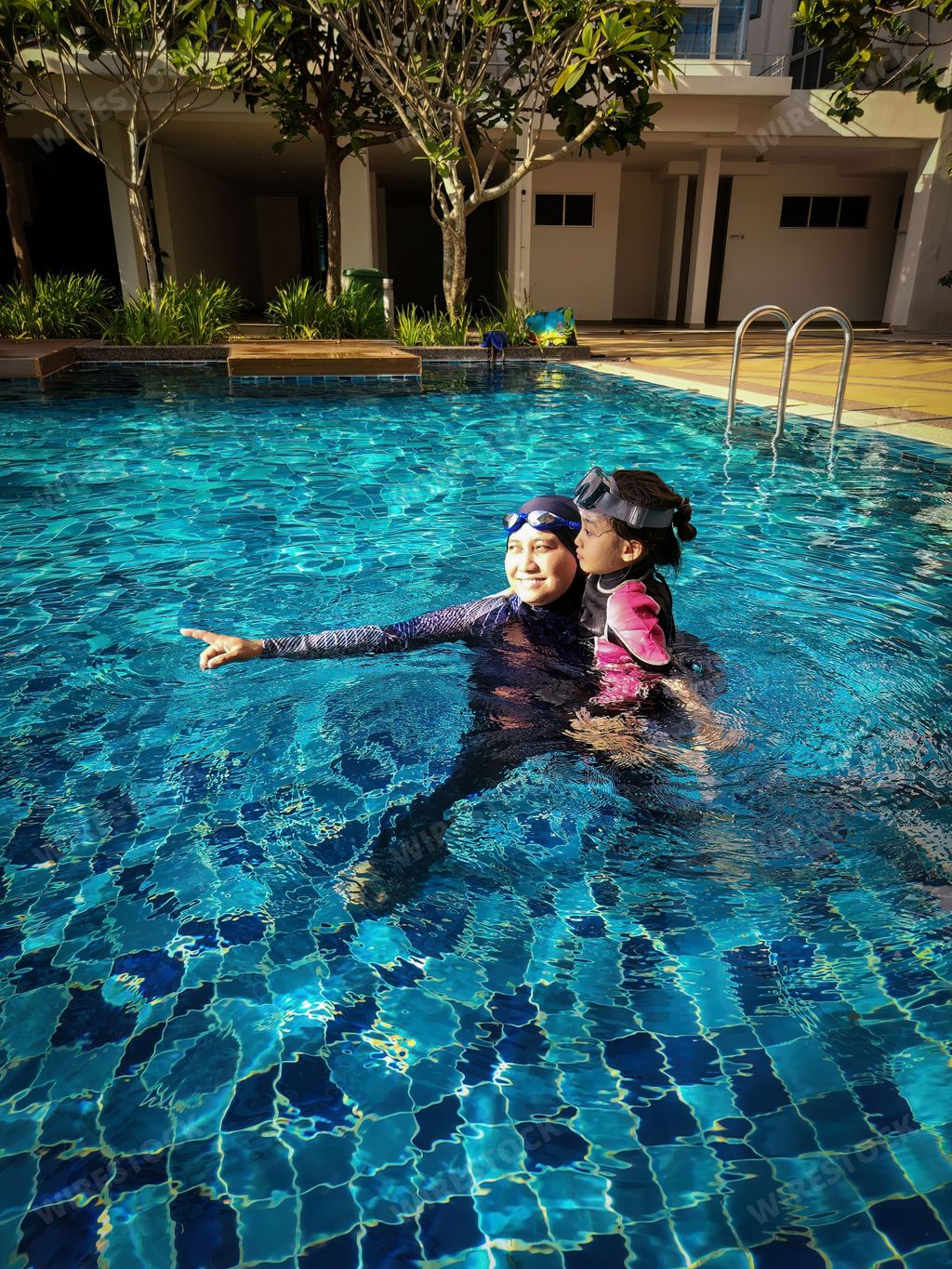 Smiling mother and her adorable little daughter hugging each other while standing together in a swimming pool at a tropical resort