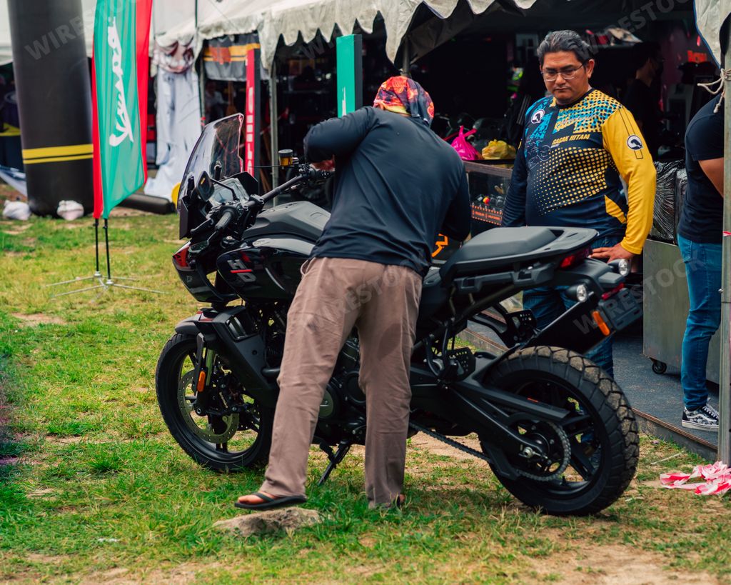 Two men looking at a black Kawasaki Versys motorcycle parked on the grass at a bike event in Terengganu