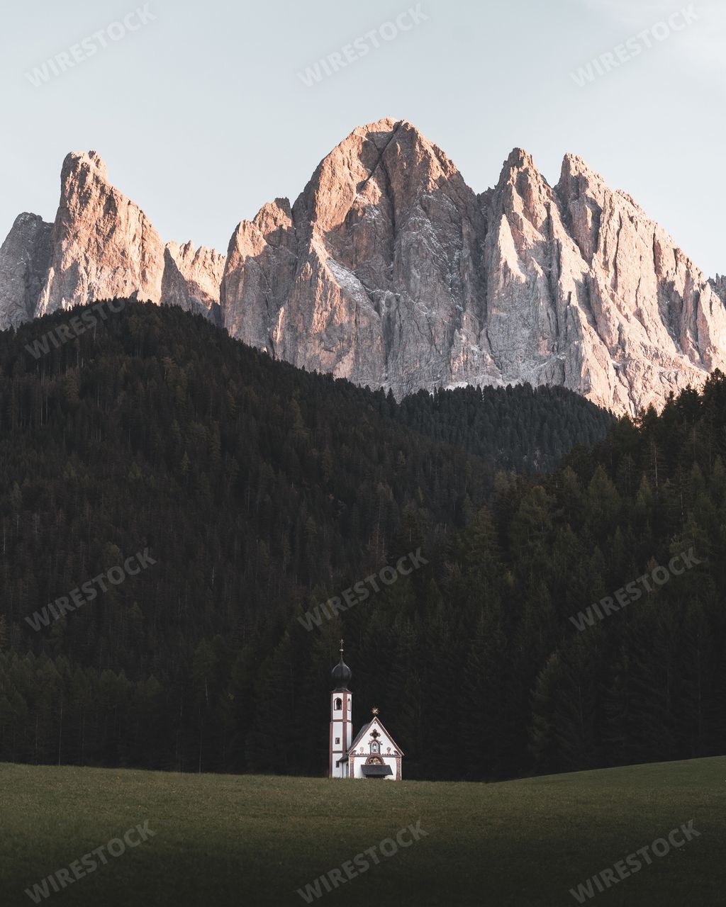 A scenic vertical shot of the church of St. John of Nepomuk in Ranui, Italy against a fir forest and rocky mountains