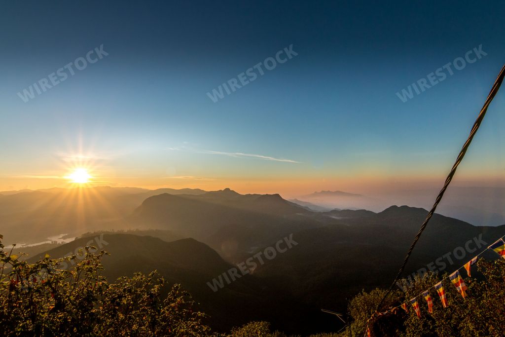 An idyllic landscape featuring the sun setting behind the majestic mountain range, visible through a cable suspension bridge