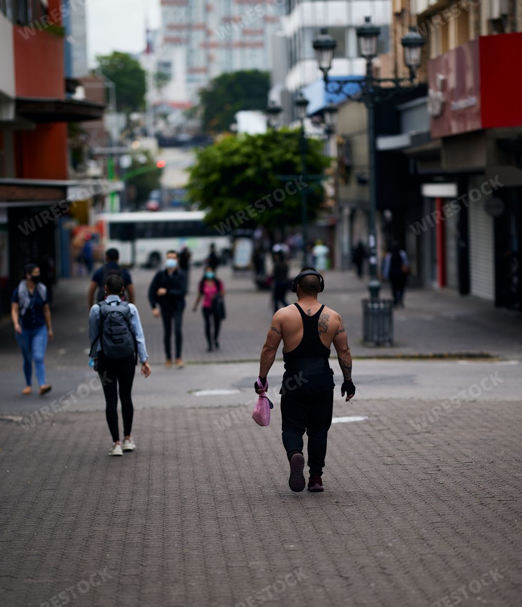 A muscular, tattooed man with headphones walking down the busy streets of San Jose during the pandemic