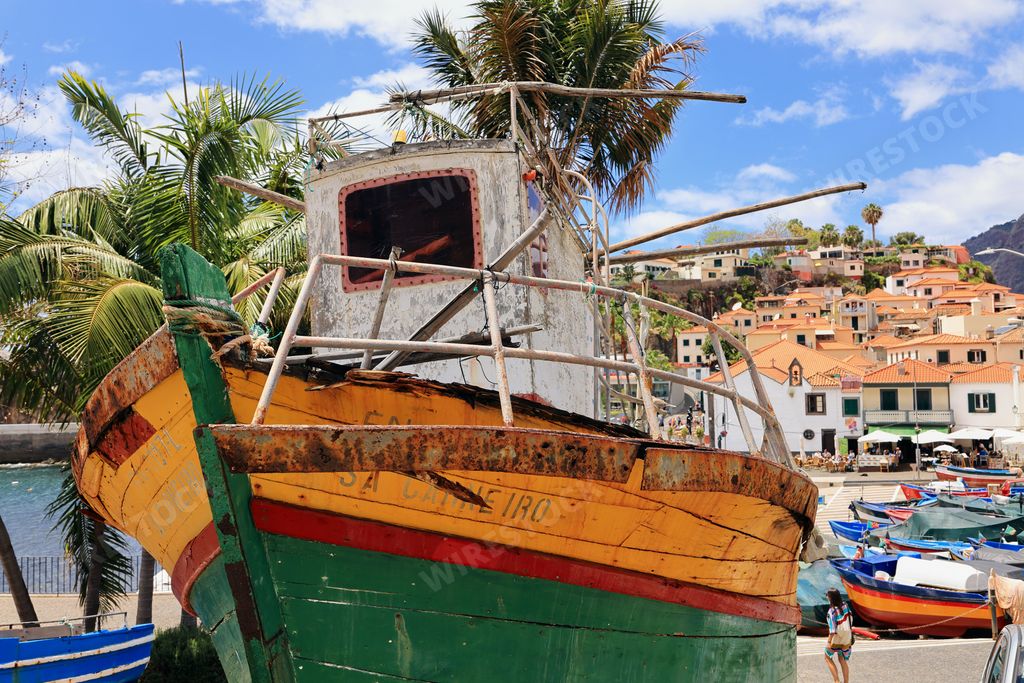An old colorful fishing boat on a sunny day in a coastal village with palm trees and houses in the background.